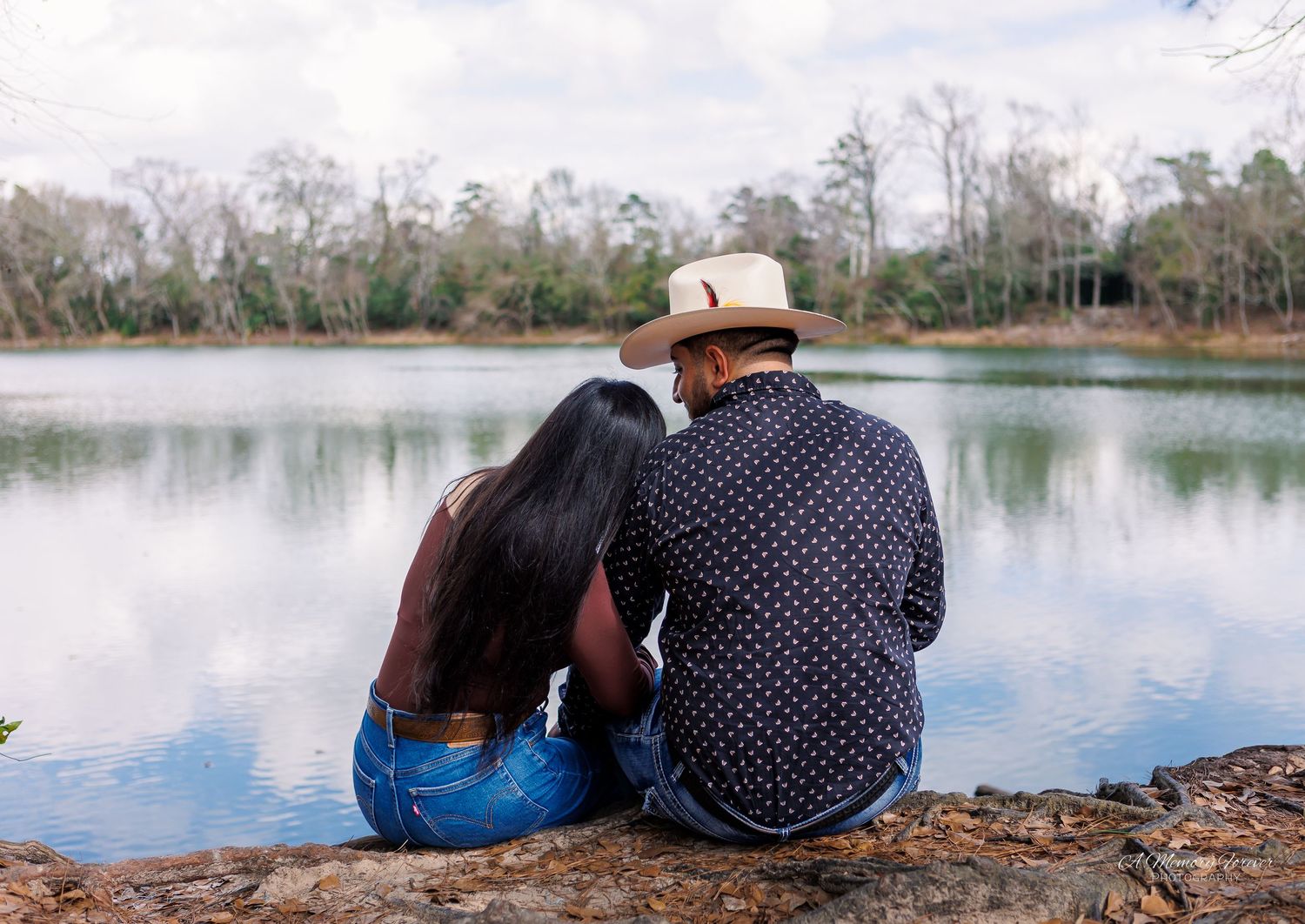 Couple sitting together on rocky lakeside, woman leaning on man wearing hat, serene water and trees in background.