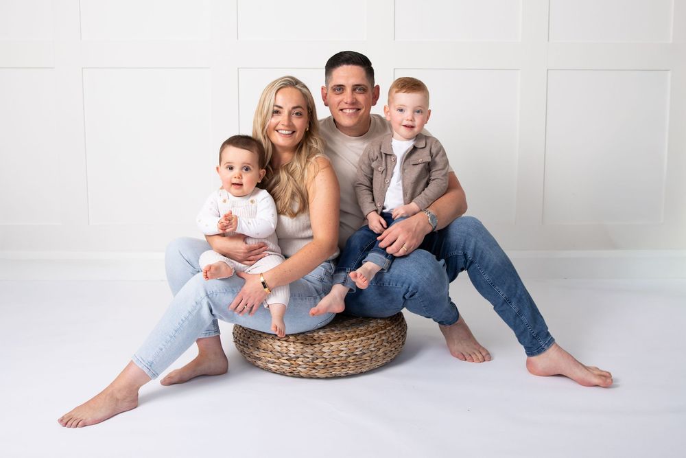 Happy family of four posing together in a bright white studio, parents sitting with two baby boys smiling.