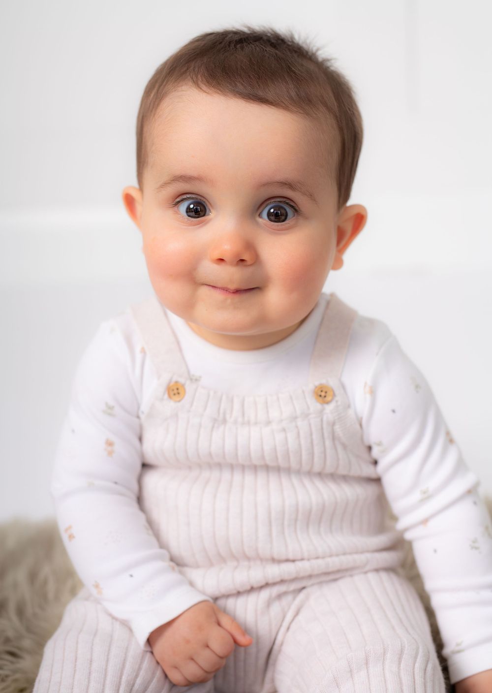 Adorable baby with big blue eyes wearing a ribbed overalls outfit and white long-sleeve shirt, smiling at the camera.
