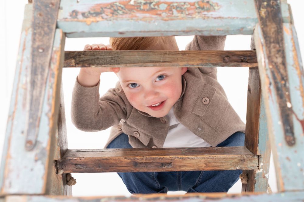 Curious toddler peeks through wooden ladder rungs, smiling playfully in a brown jacket and jeans.