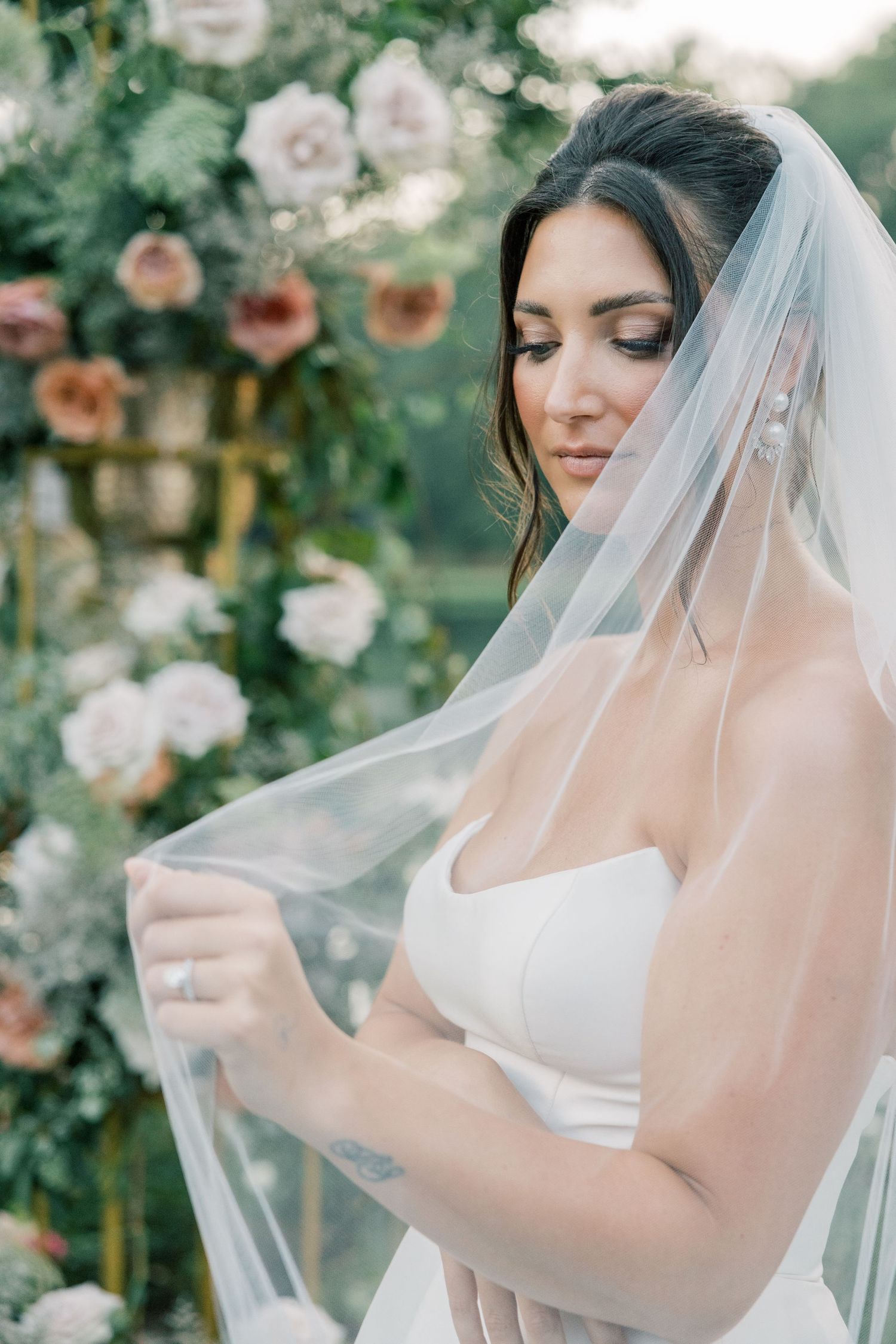 Elegant bride in strapless gown holding sheer veil with floral arch backdrop outdoors.