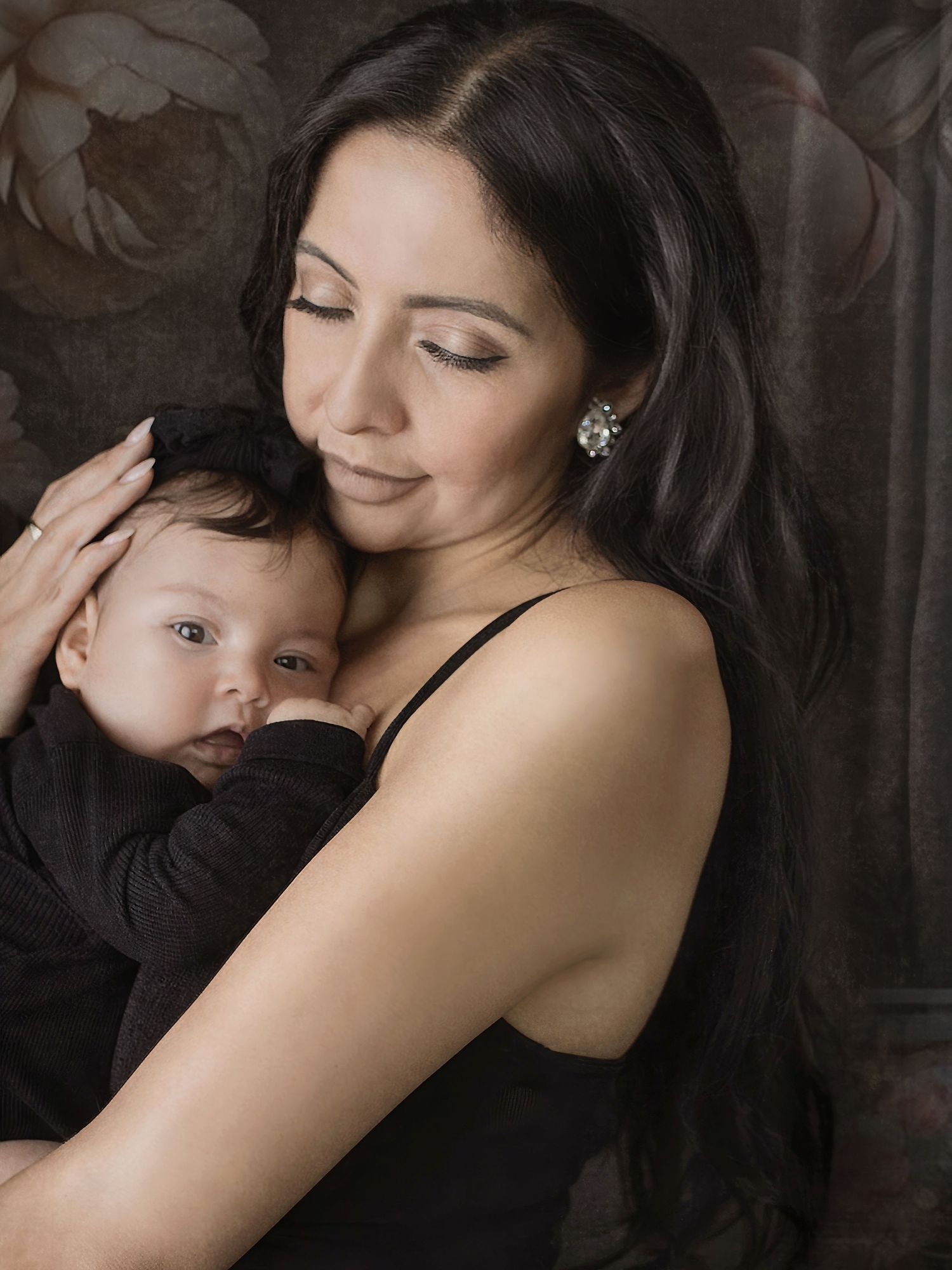 A tender moment as a parent in a black top embraces a small baby with dark hair against a moody brown background.