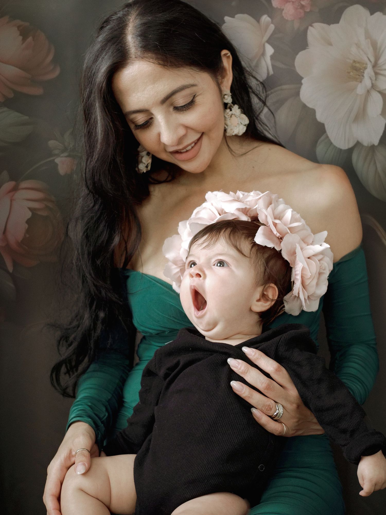 Mother in emerald green dress shares tender moments with baby in black outfit against floral backdrop.