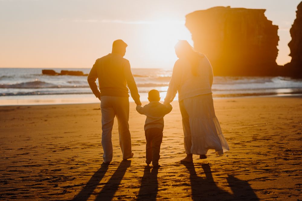 Three silhouettes walking on a beach at sunset with a large rock formation in the background.