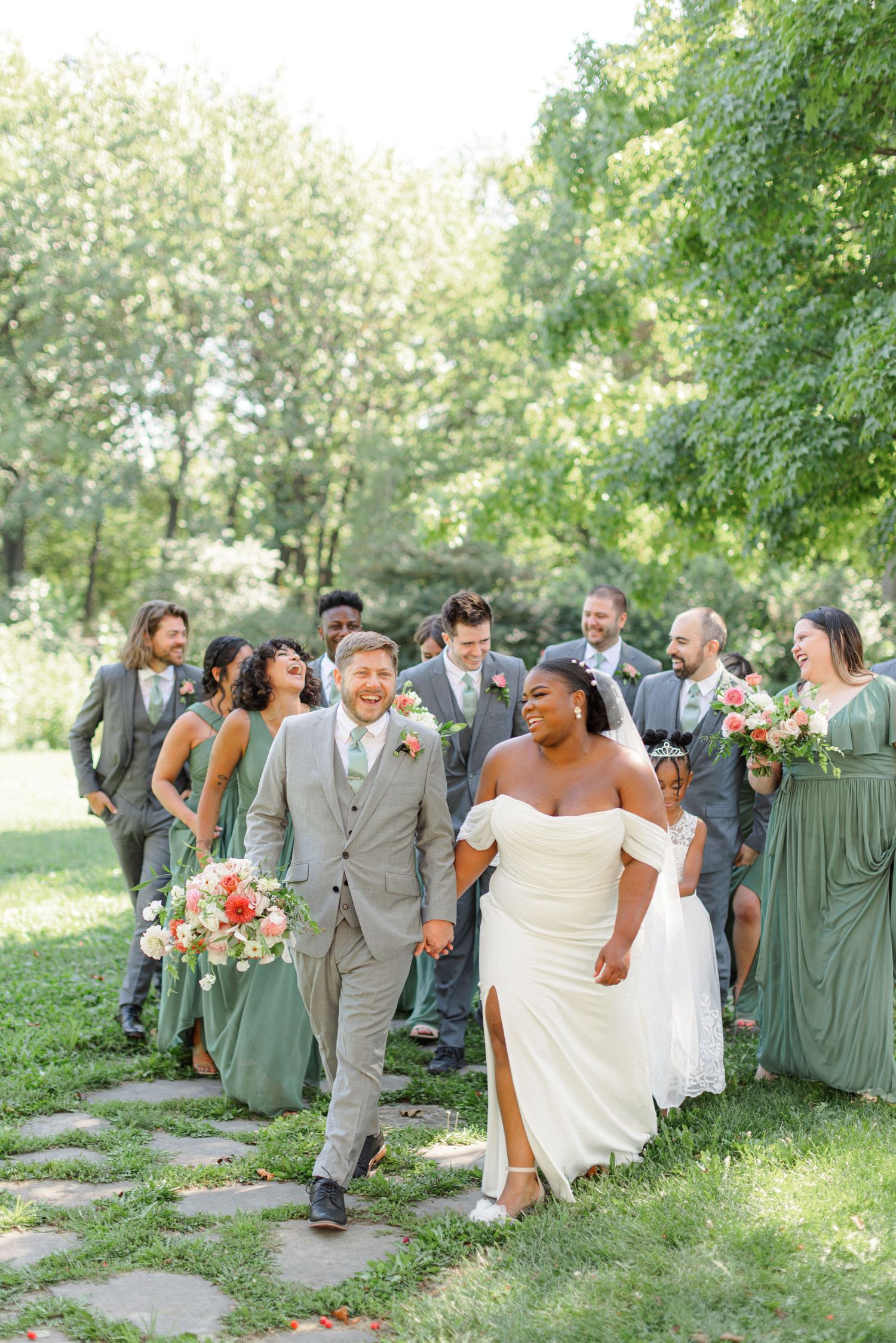 Wedding party walking along a garden path in sage green attire on a sunny day.