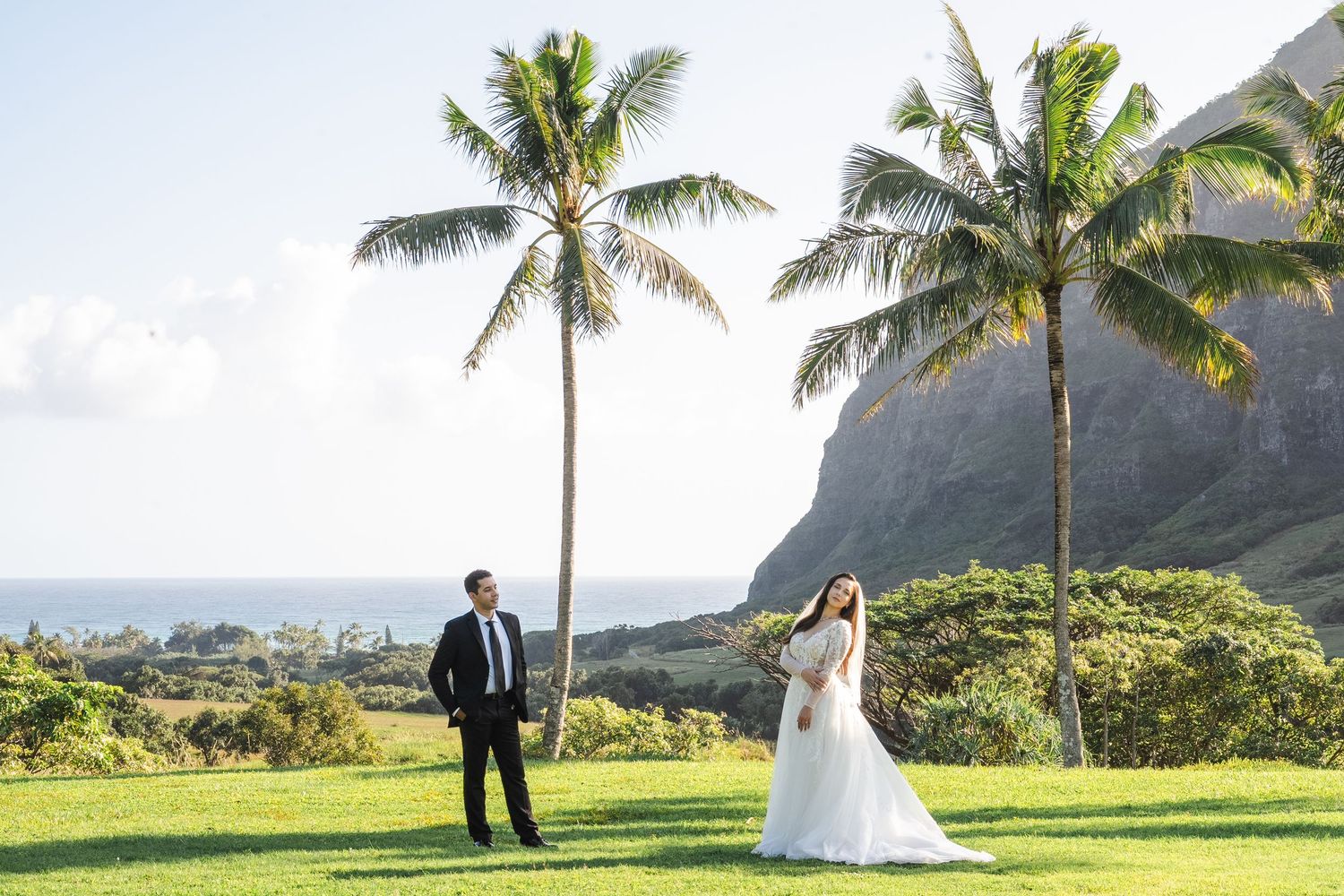 Couple in formal attire stands together on a lush green field below dramatic mountain peaks at Kualoa Ranch, Hawaii.