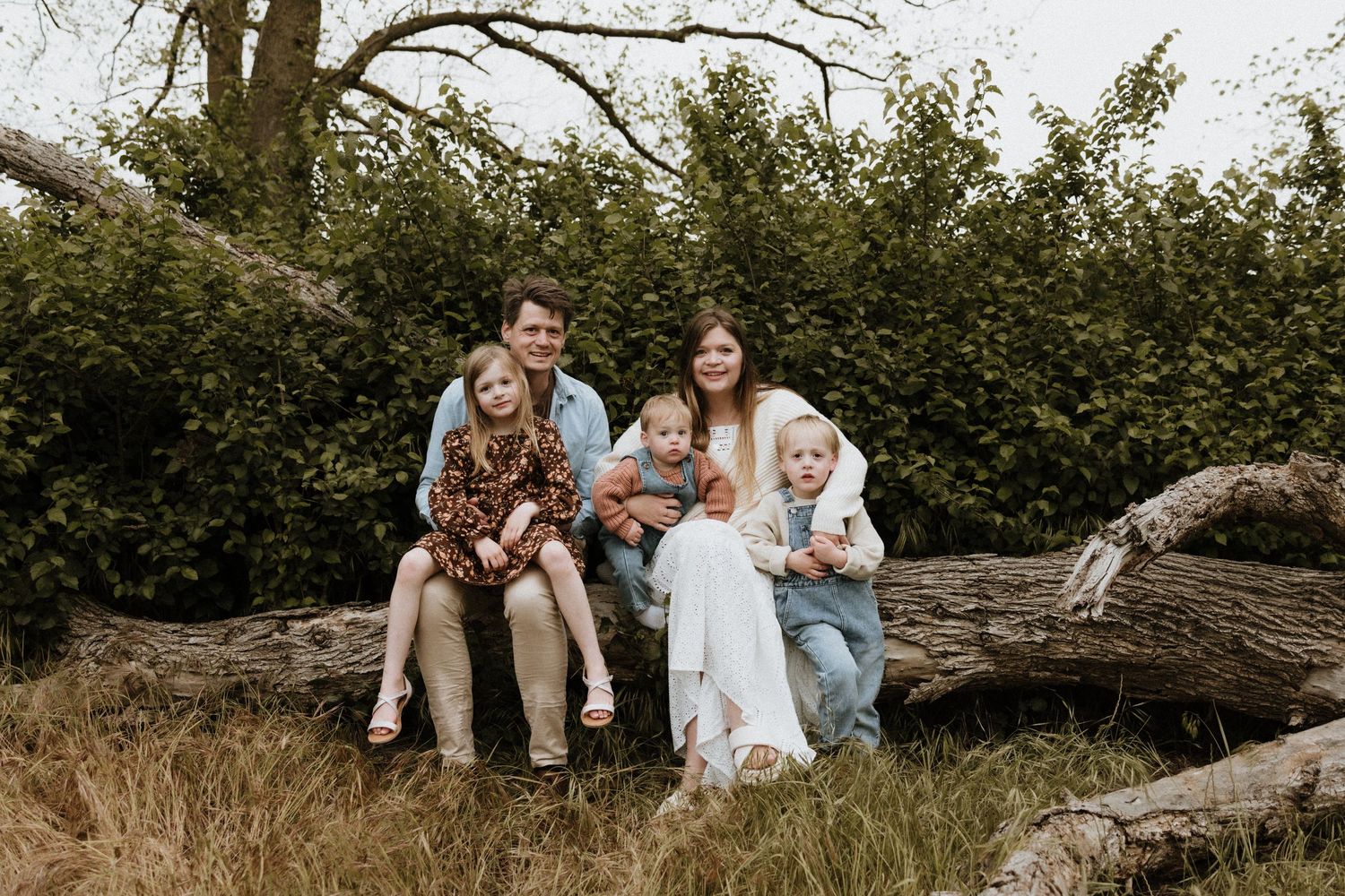 A family of five sitting together on a fallen log in a lush green outdoor setting.