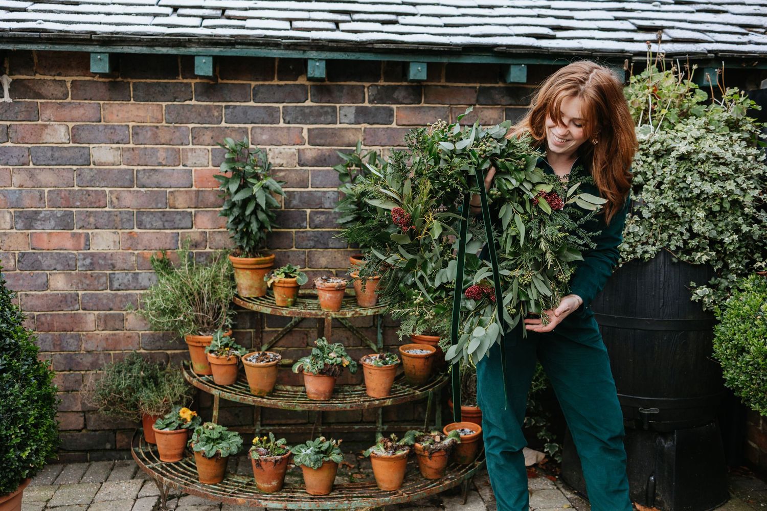 A gardener in green overalls tends to a tiered display of terracotta potted plants against a brick wall.