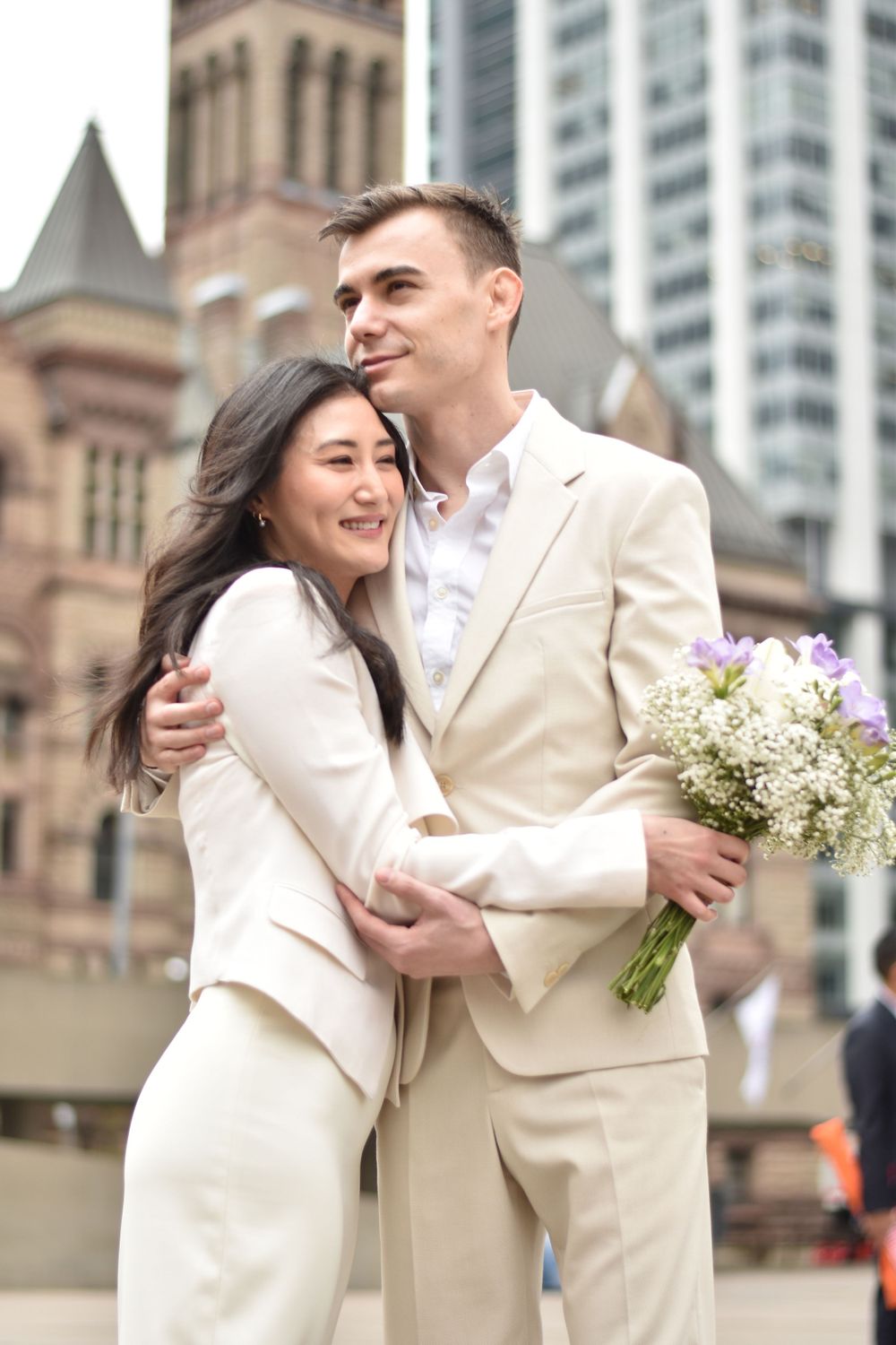 Happy couple in matching cream suits embracing with a bouquet in the city. Toronto GTA