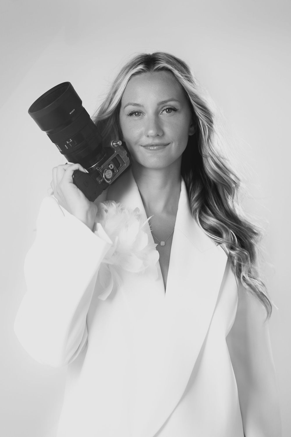 Black and white portrait of blonde female photographer holding DSLR camera wearing white blazer jacket.