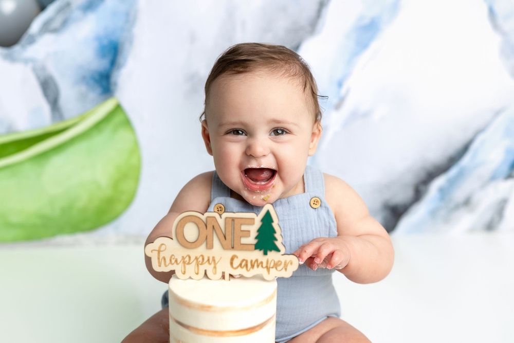 Smiling baby boy in blue romper celebrating first birthday with ONE Happy Camper cake topper on white cake.