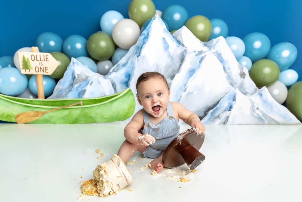 Baby boy smashes first birthday cake surrounded by mountain backdrop, balloons, and canoe at an outdoor adventure themed party.