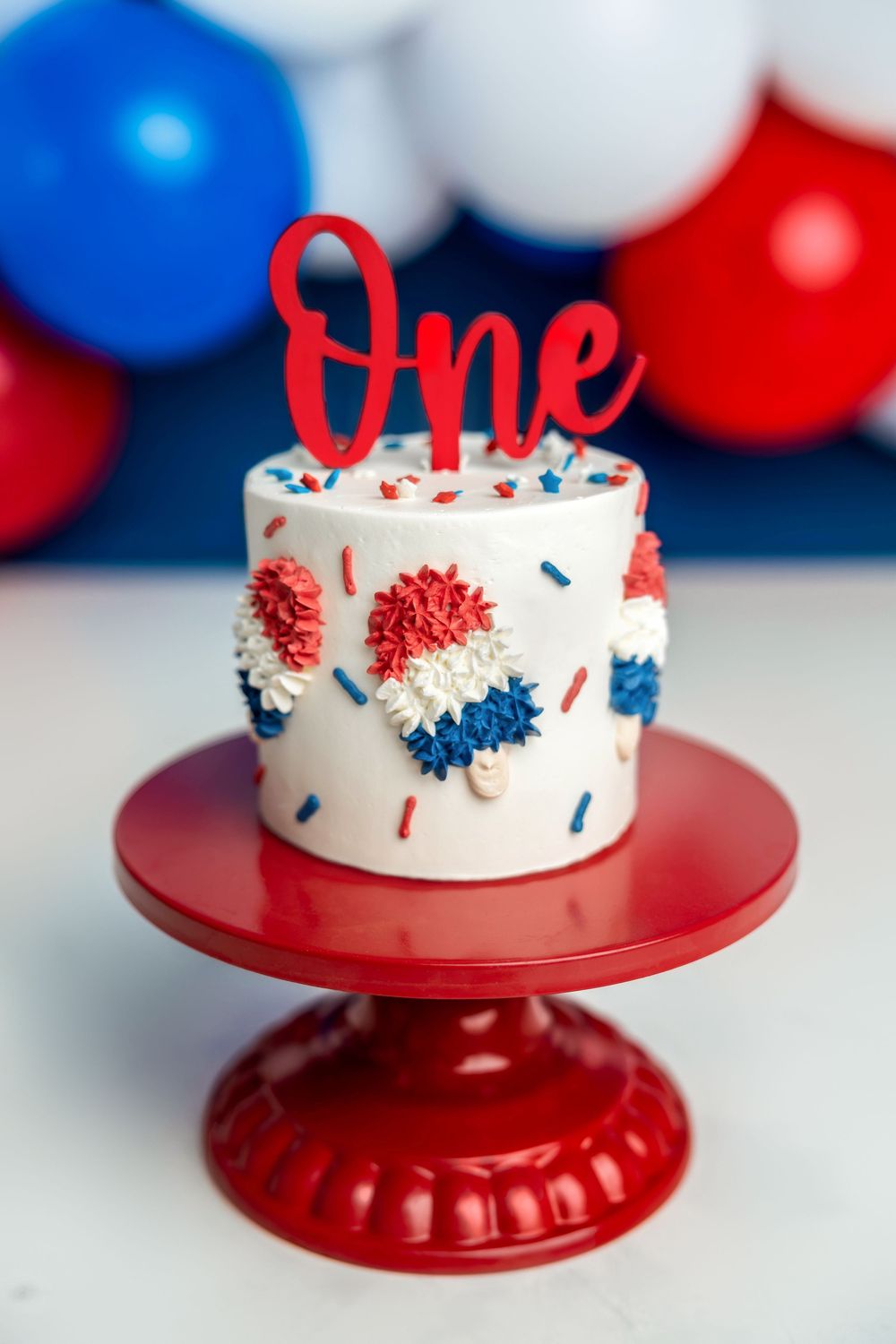 Patriotic first birthday smash cake with red, white, blue frosting and sprinkles on red cake stand with balloon backdrop.