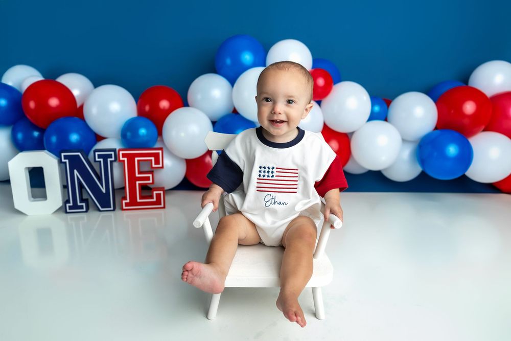 Baby boy in American flag onesie sits smiling at his first birthday party with red, white, and blue balloons.