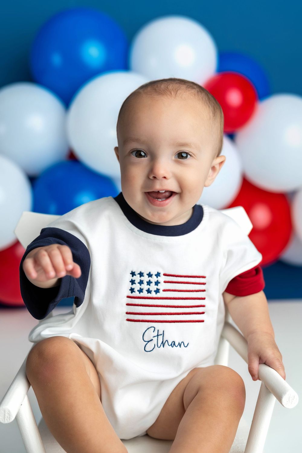 Smiling baby named Ethan wearing patriotic American flag romper sits in white chair with red, white, and blue balloons.