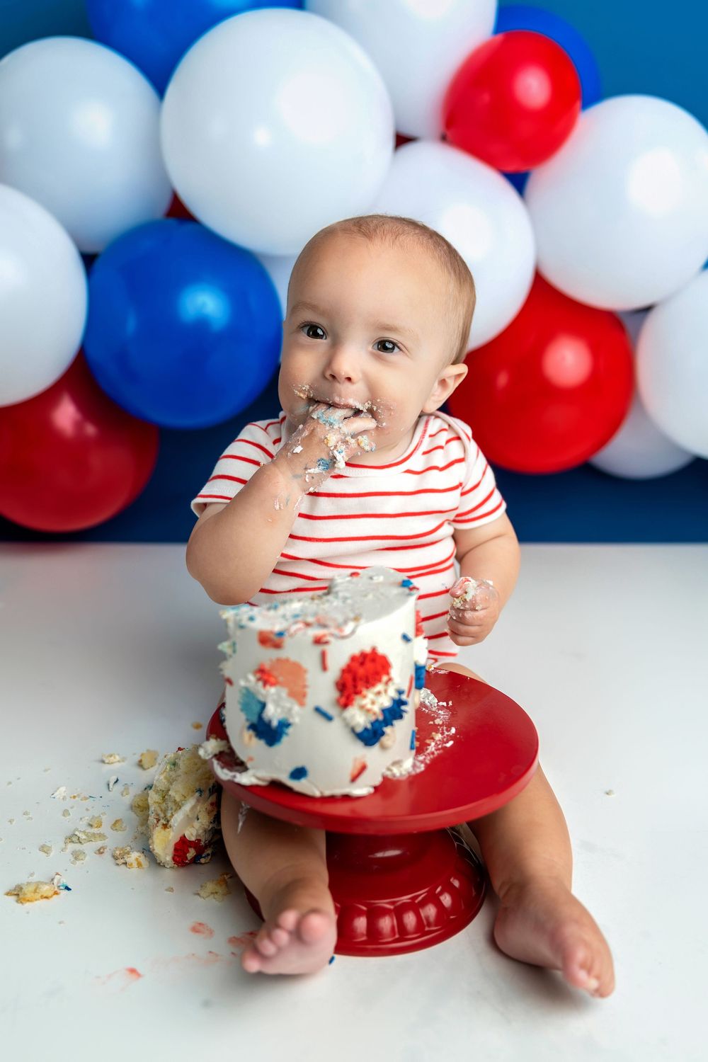 Baby eating first birthday cake during a patriotic red, white, and blue cake smash photoshoot with balloons.