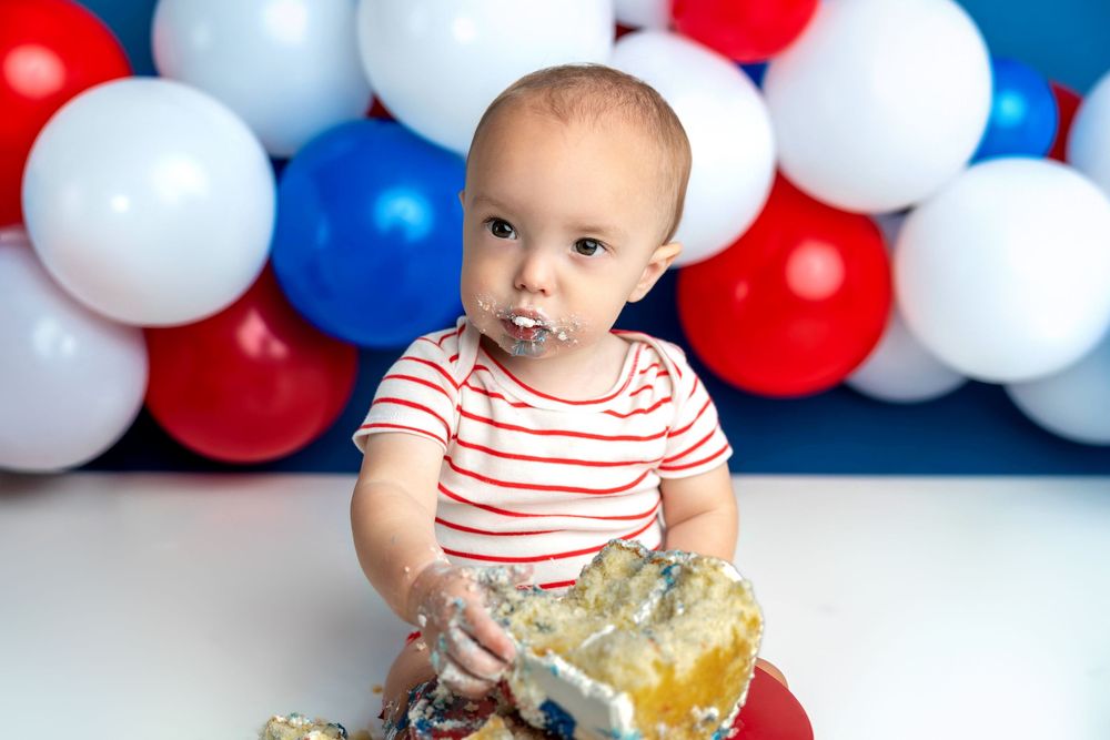 Baby with cake smeared on face enjoys first birthday cake smash with red, white, and blue balloon backdrop.
