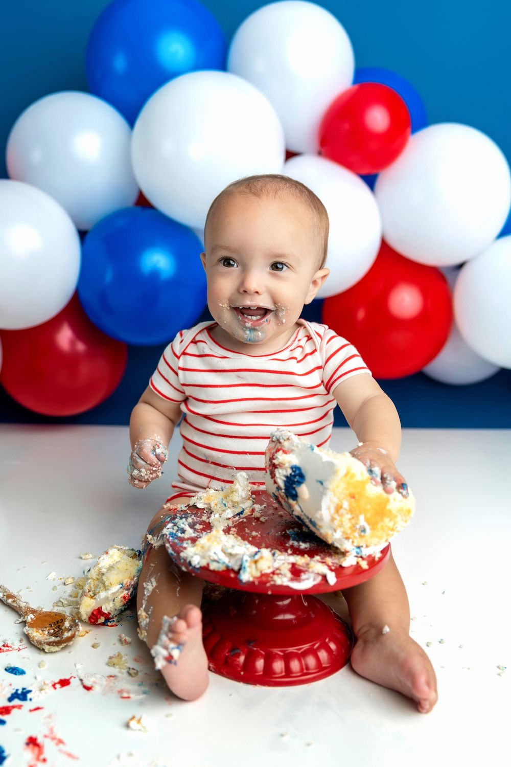 Baby smashing first birthday cake on red stand with red, white, and blue balloons in patriotic cake smash photo session.