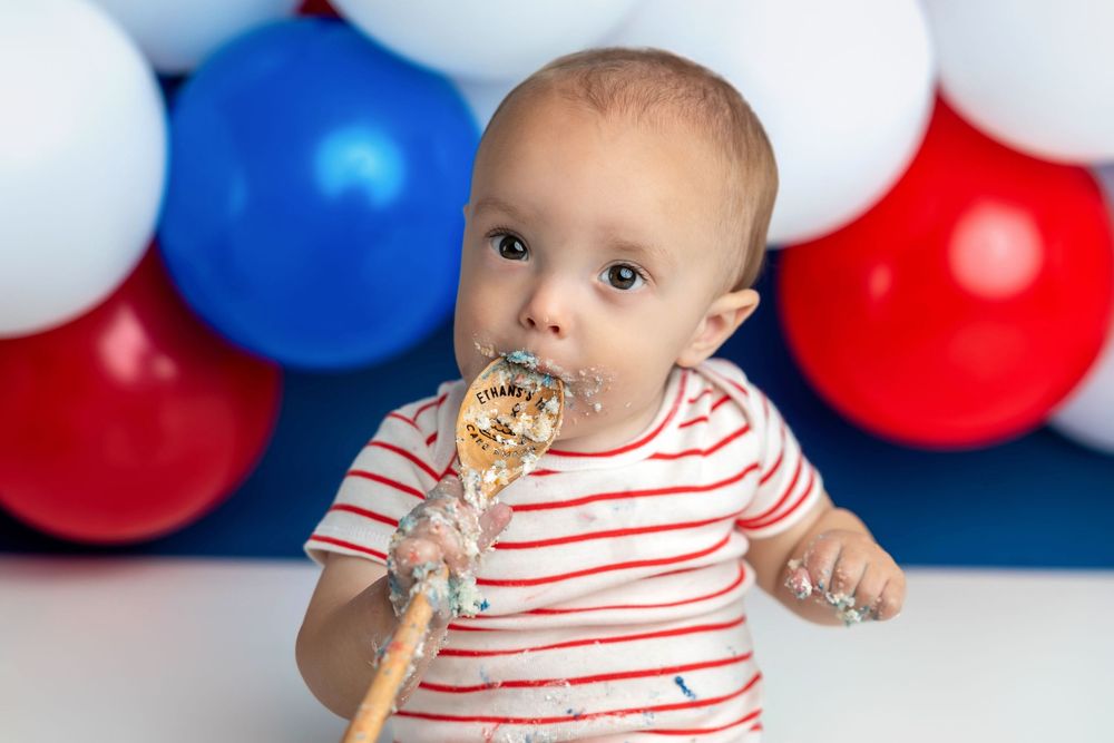 Baby in red striped shirt eating cake smash with colorful red, white, and blue balloons in background.