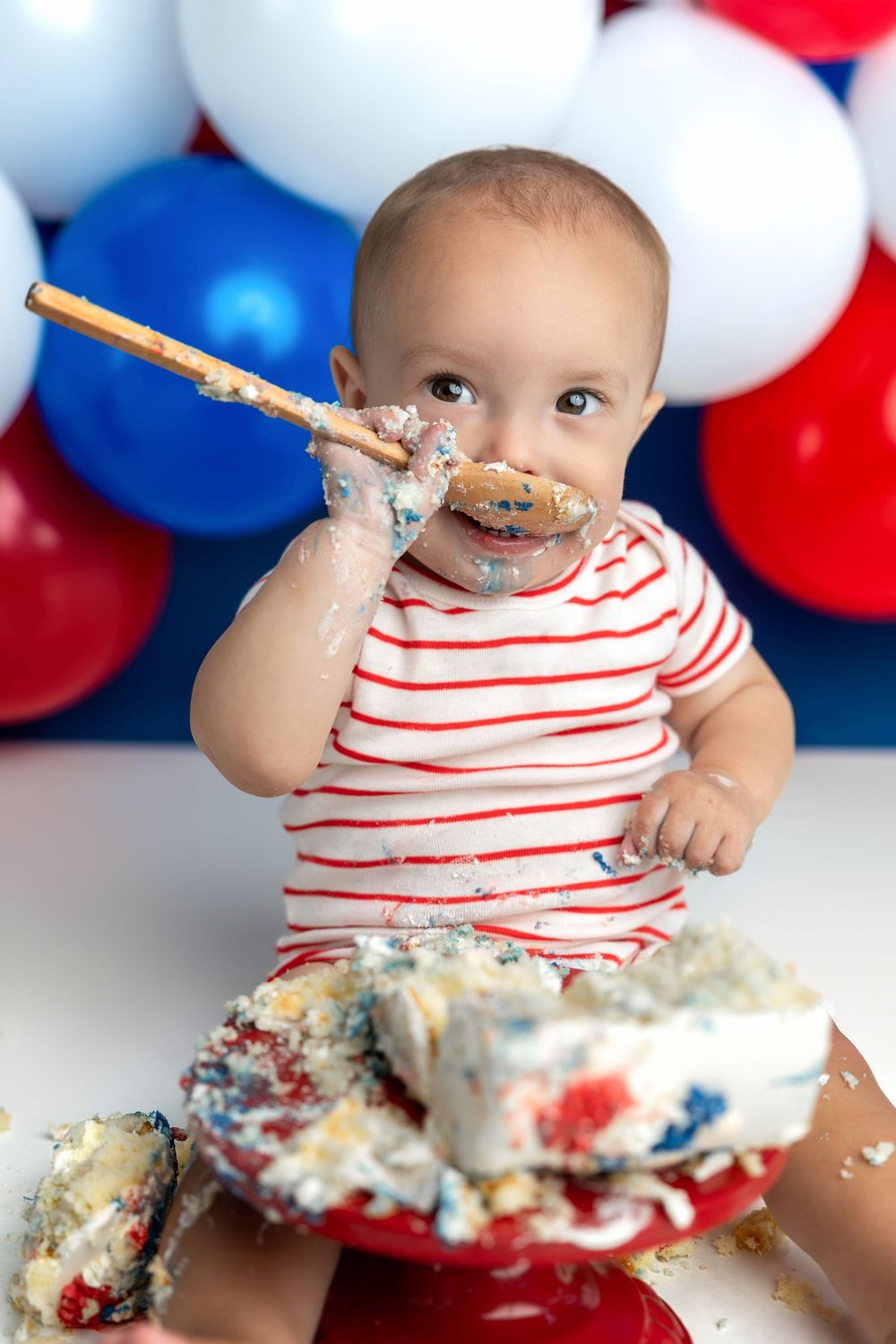 Baby in red striped shirt smashing first birthday cake with red, white, and blue balloons in background.
