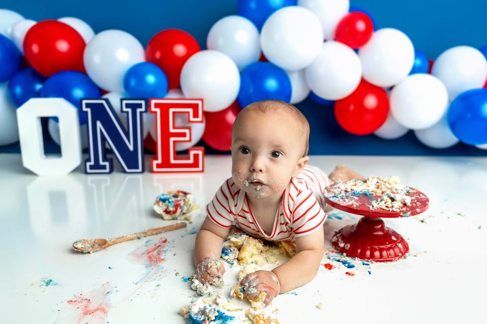 Baby's first birthday cake smash with red, white, and blue balloons, ONE letters, and smashed cake on white floor.