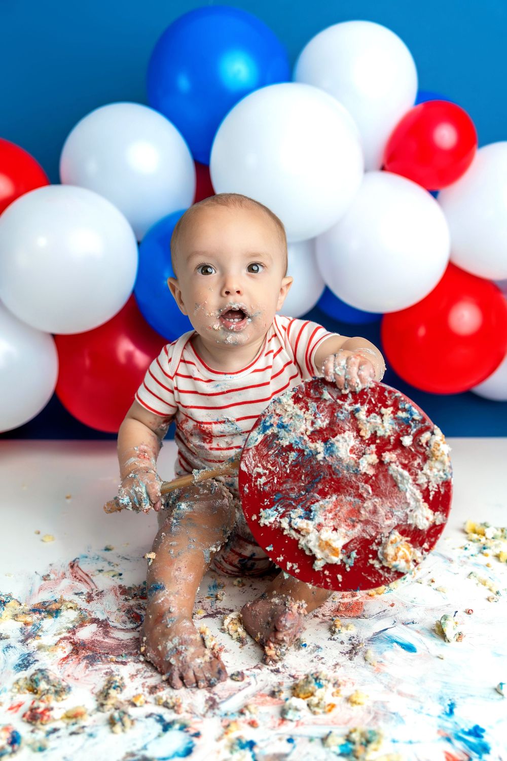 Messy baby smashes red birthday cake during patriotic cake smash session with colorful balloon garland behind.