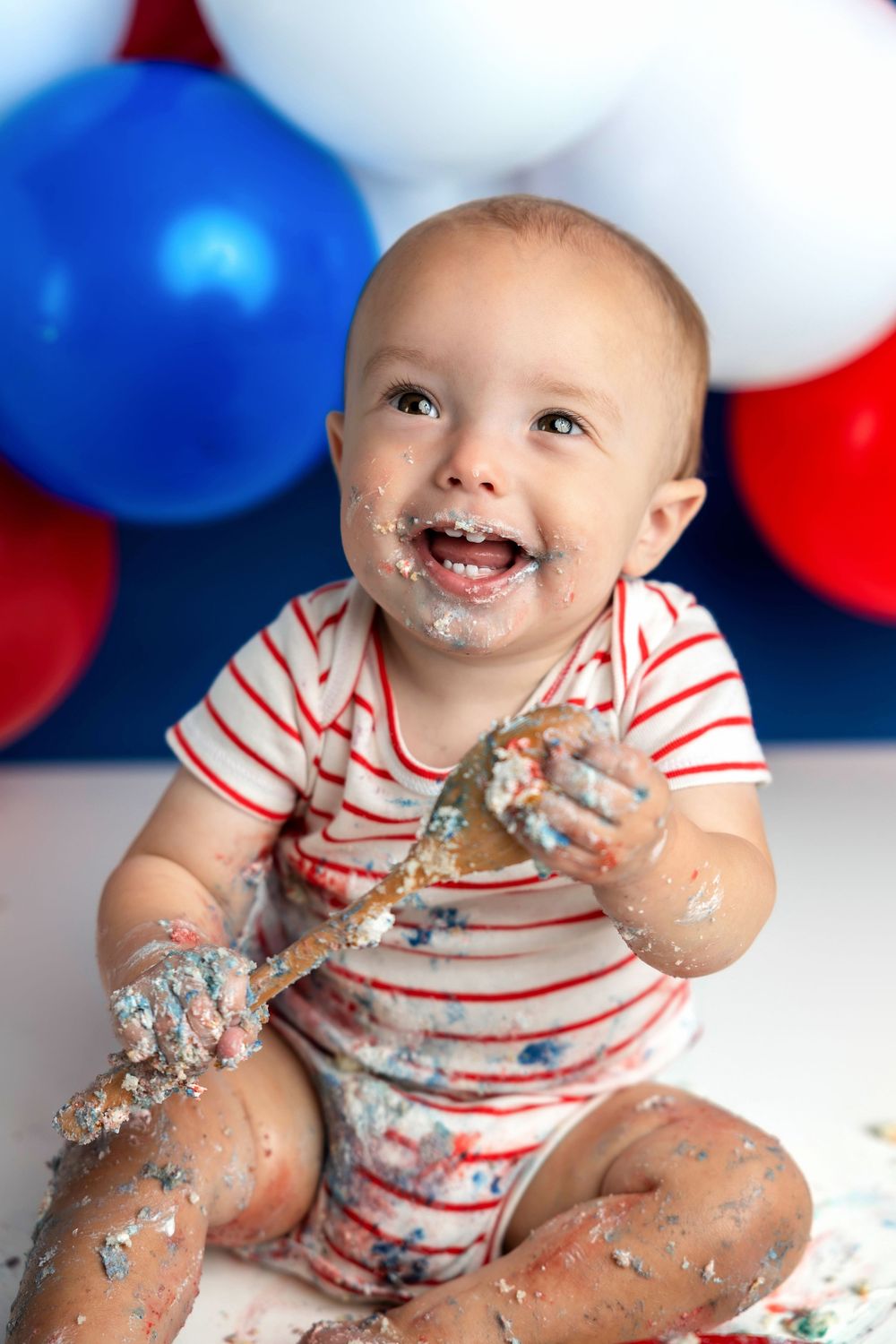 Happy baby covered in cake during first birthday smash cake session with red, white, and blue balloons.