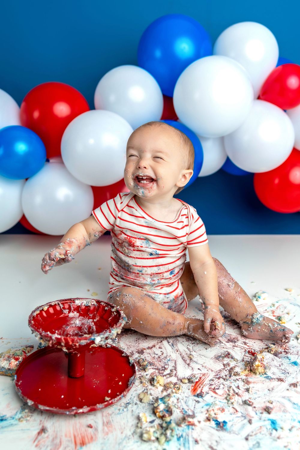Laughing baby covered in cake during a patriotic red, white, and blue first birthday cake smash photoshoot.