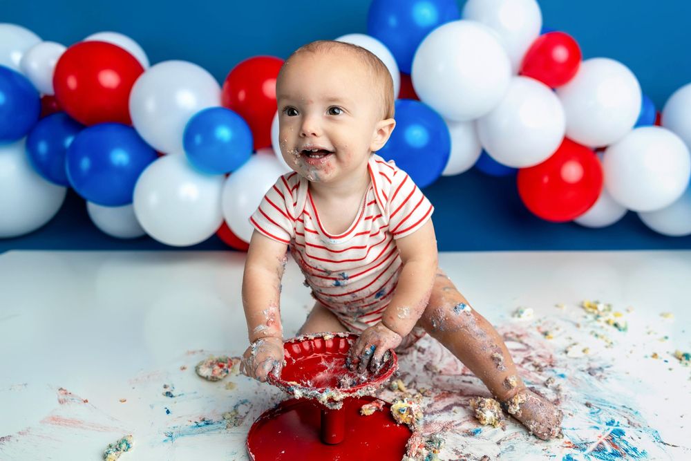 Happy baby smashes red cake at patriotic first birthday party surrounded by red, white, and blue balloons.