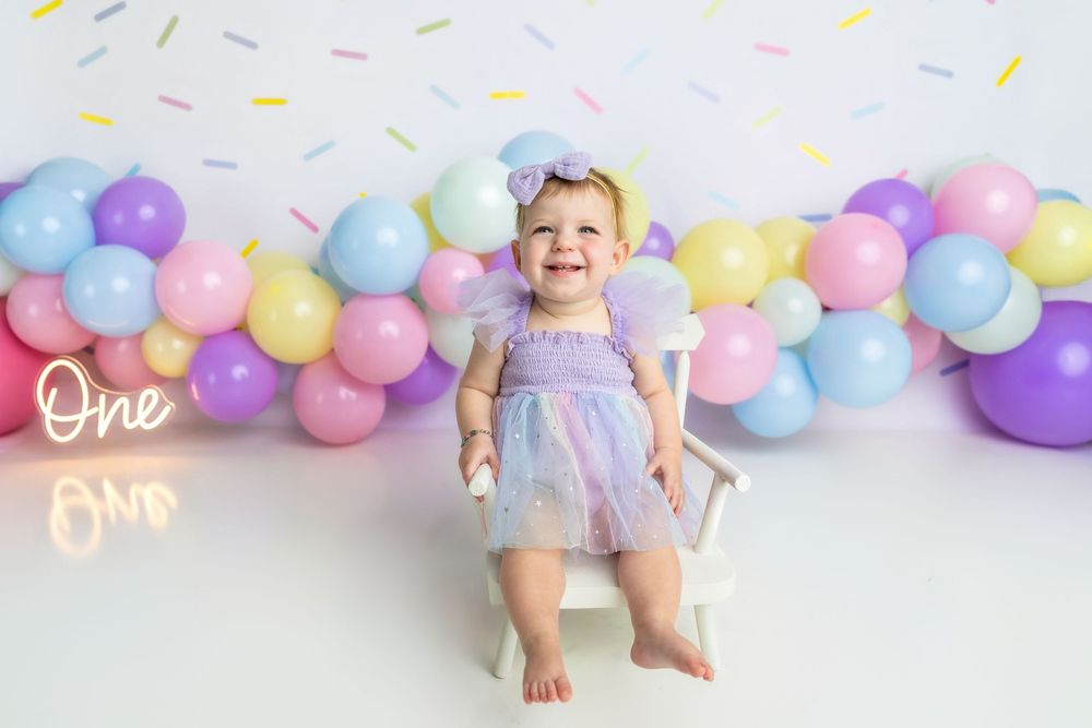 Baby girl in purple tutu sits smiling among pastel balloons for a first birthday cake smash photo.