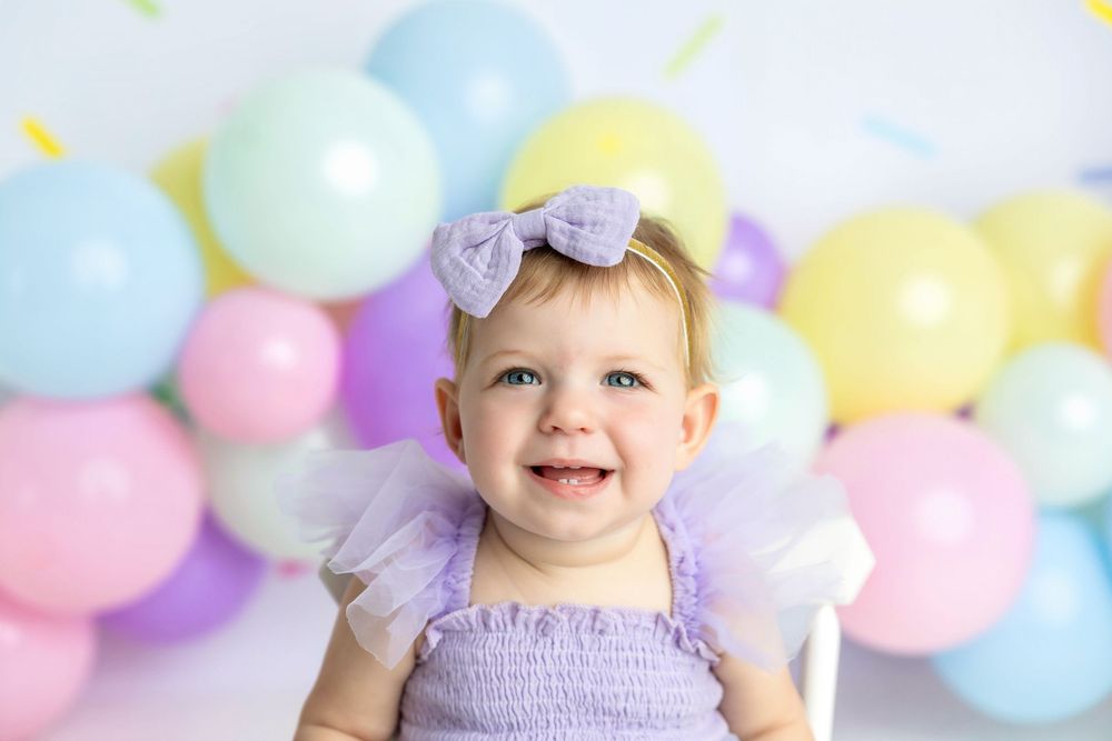 Smiling baby girl in purple outfit with bow headband at colorful balloon birthday party photo shoot.