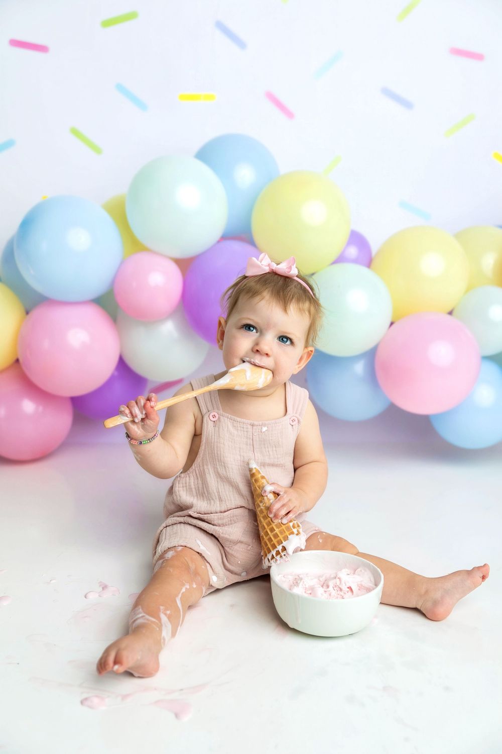 Baby girl in romper eating cake with wooden spoon at colorful balloon birthday smash cake photoshoot.