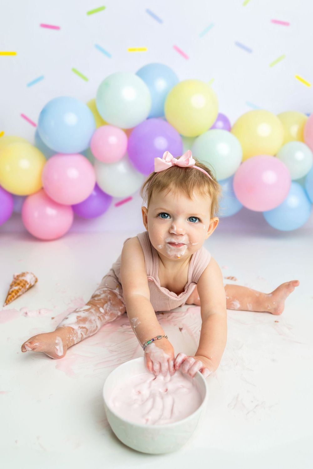 Baby girl in pink dress smashes first birthday cake, surrounded by colorful pastel balloons on white background.