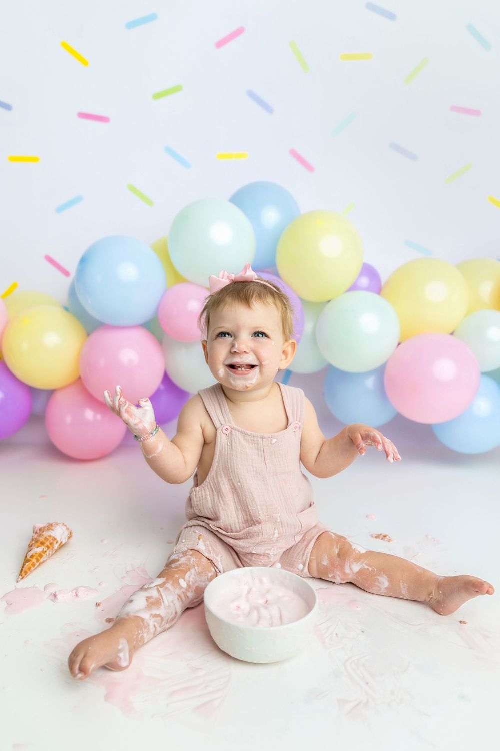Baby girl in pink romper smashes first birthday cake, sitting among colorful balloons on white backdrop.