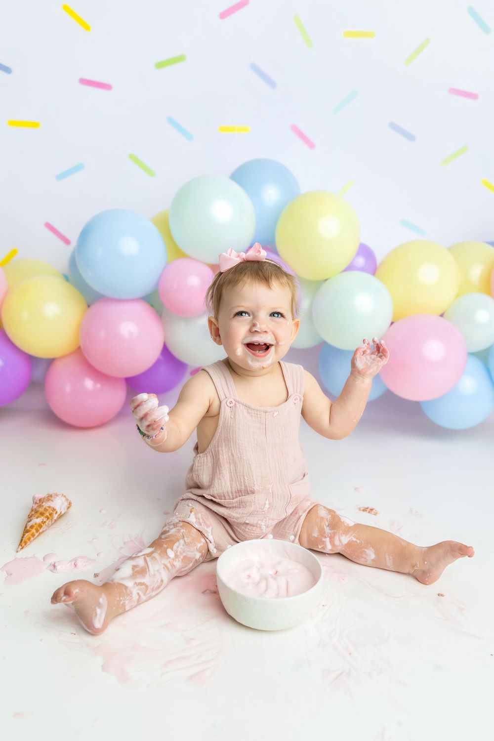 Baby girl in pink romper smashes first birthday cake, surrounded by pastel balloons and colorful sprinkle backdrop.
