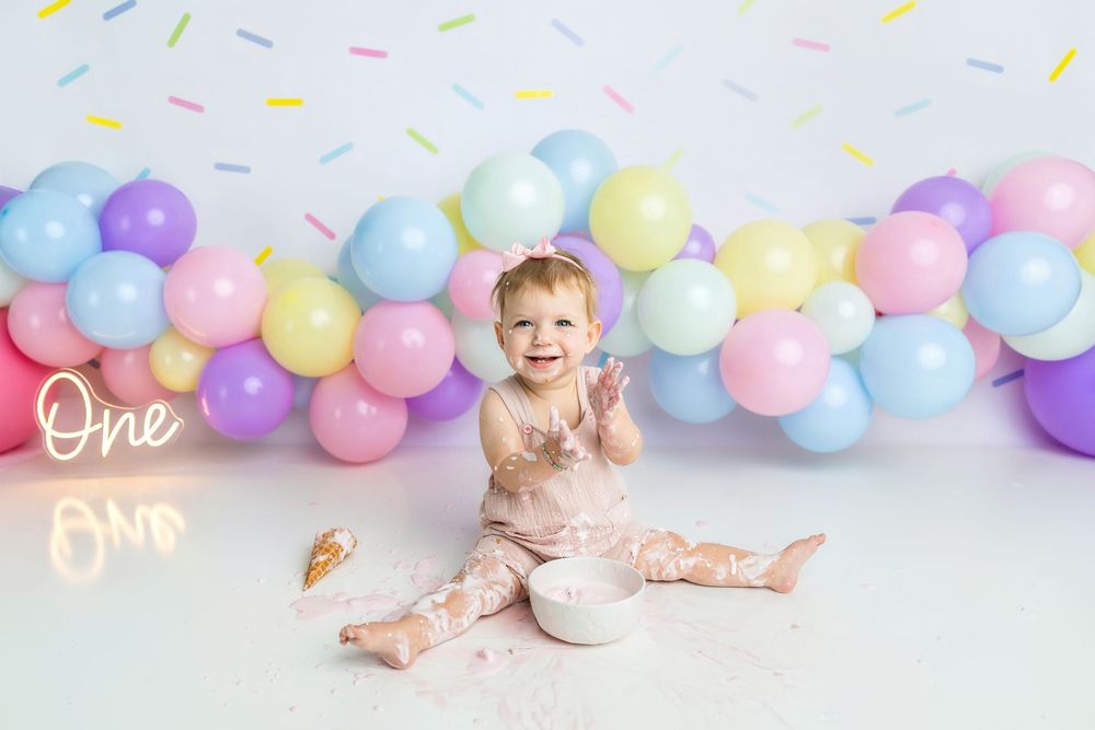 Baby girl smashes first birthday cake surrounded by pastel balloons and a glowing 'One' sign.