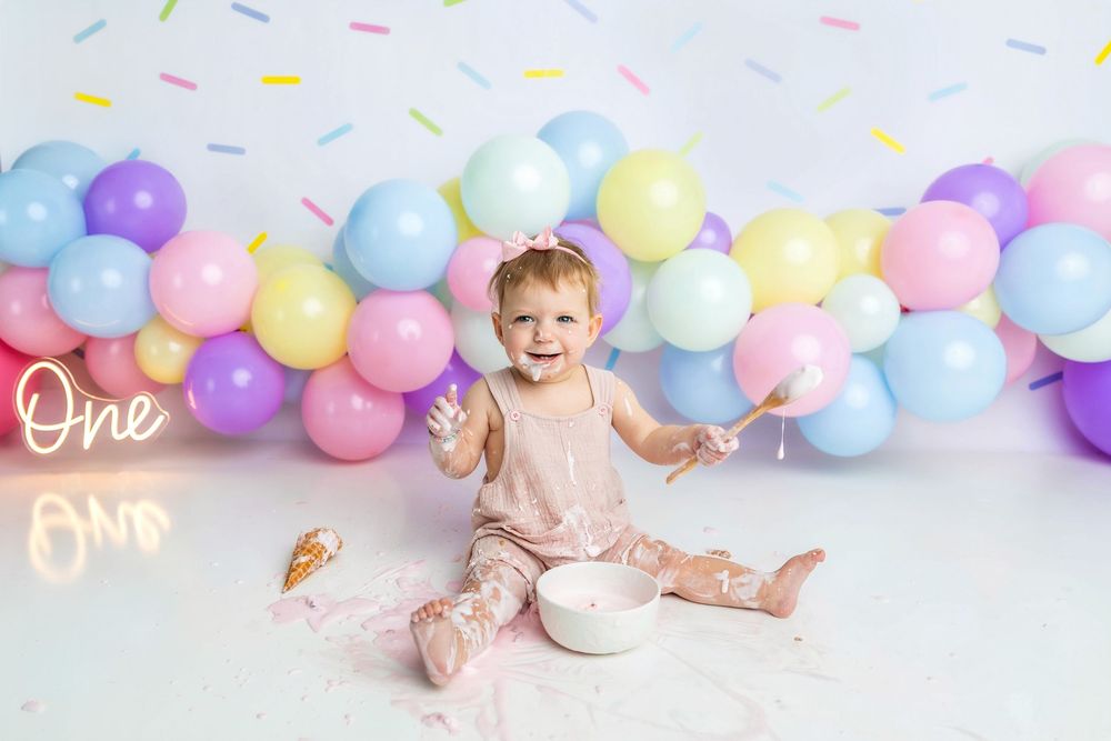 Baby girl celebrates first birthday smashing cake surrounded by pastel balloons and a glowing 'One' sign.