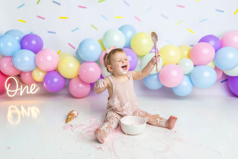 Baby girl laughing during first birthday cake smash with colorful balloon garland and neon One sign.