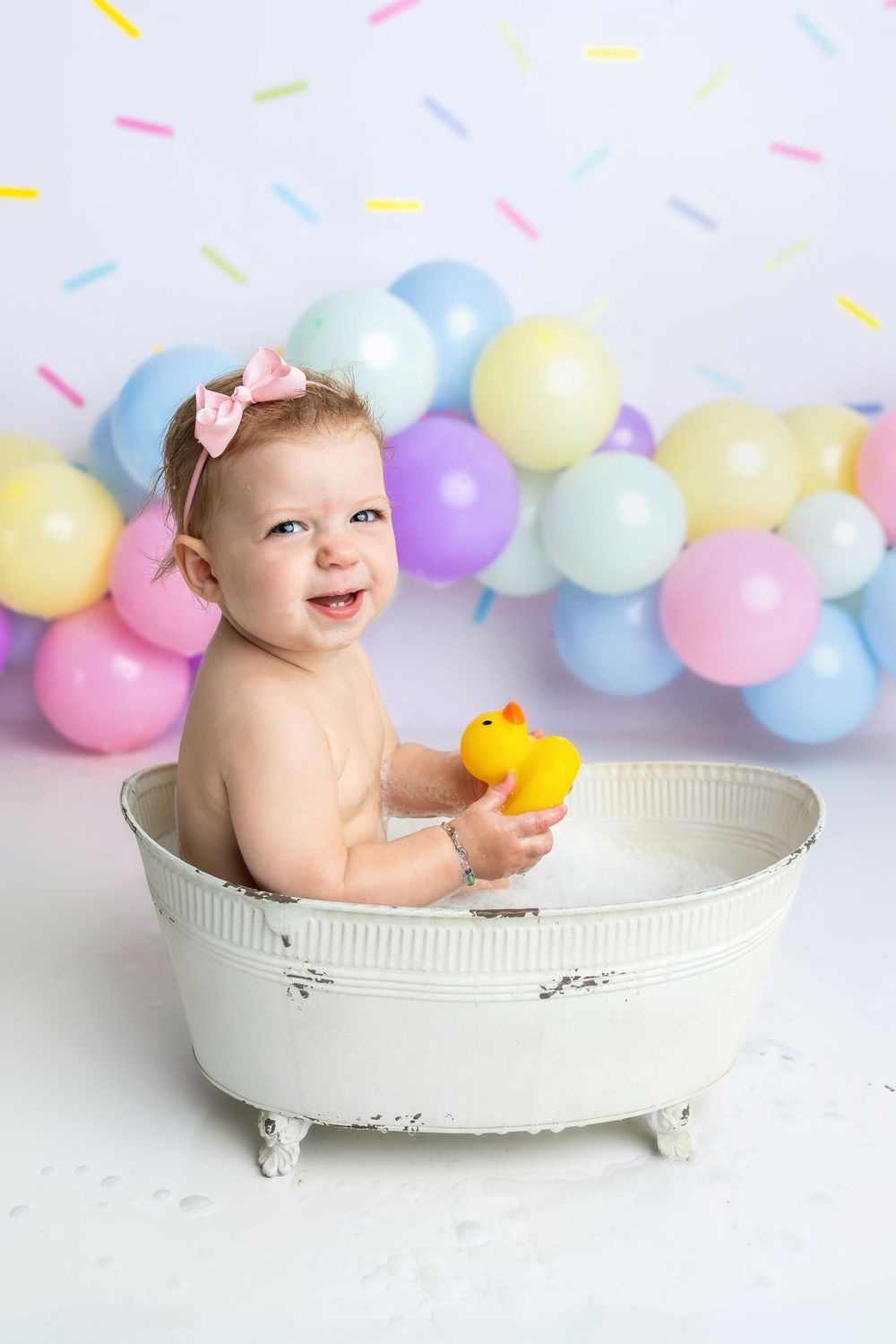 Smiling baby girl in a white mini bathtub holding a rubber duck, surrounded by colorful balloons and confetti.