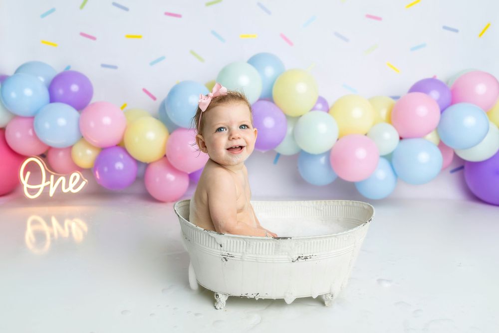 Baby girl smiling in white vintage tub at first birthday cake smash with colorful balloon garland backdrop.