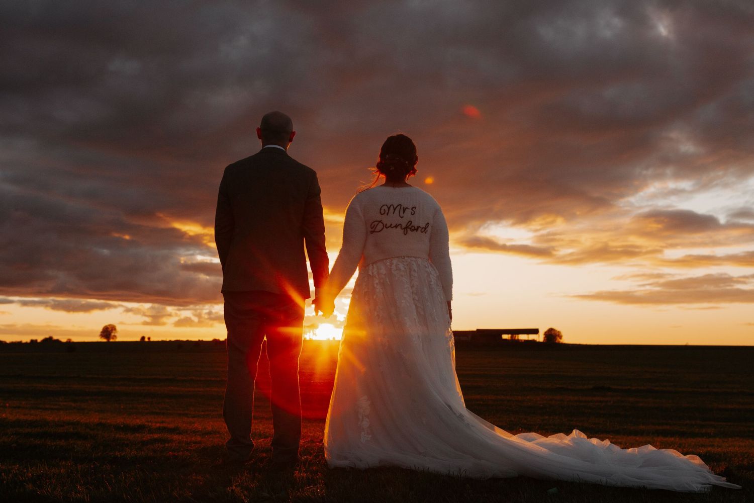 Bride and groom holding hands at sunset in open field, bride wearing custom embroidered wedding gown with dramatic cloudy sky.