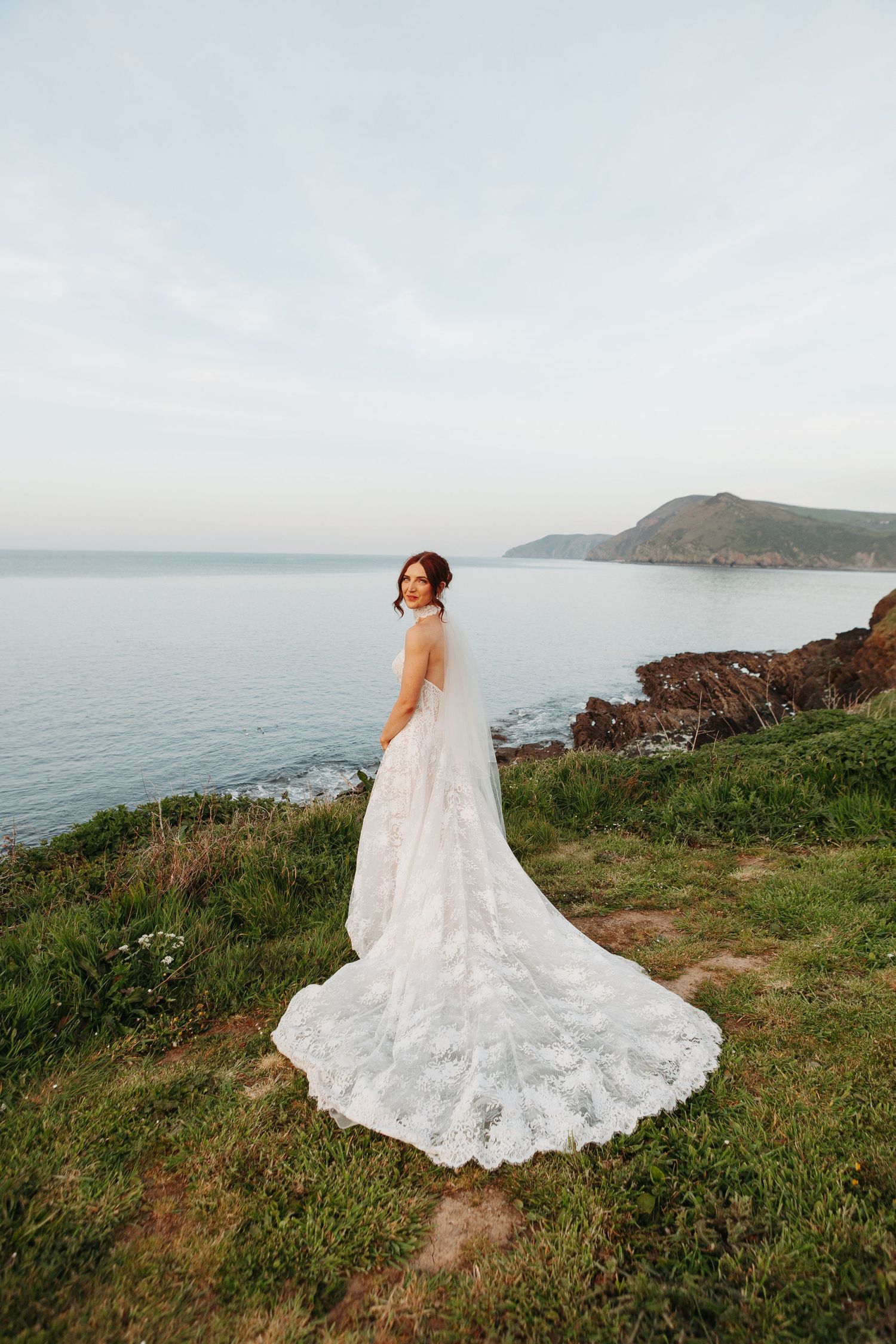 Bride in lace wedding gown with long train stands on coastal cliff overlooking serene ocean and green hills.