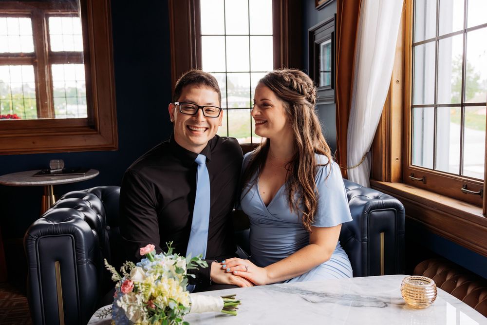 Happy couple sitting together at a white table in an elegant dark-wood restaurant, smiling and holding hands on their wedding day.