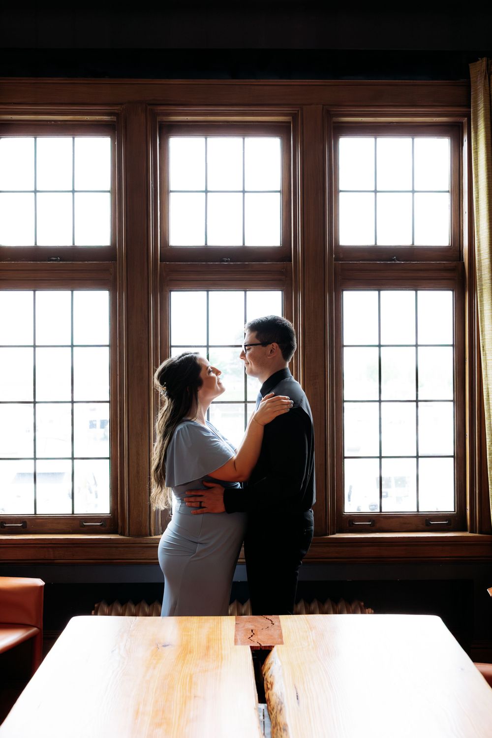 Couple embracing romantically by large wooden-framed windows in an elegant indoor setting with warm lighting.