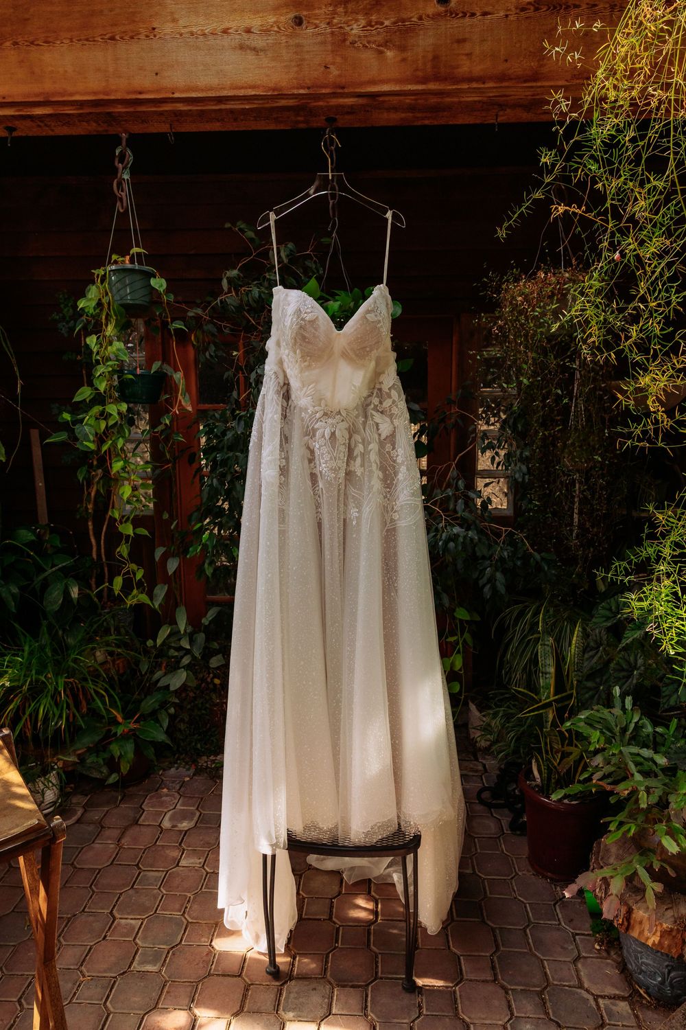 Elegant white lace wedding dress hanging on a hanger outdoors, surrounded by lush green plants and wooden backdrop.