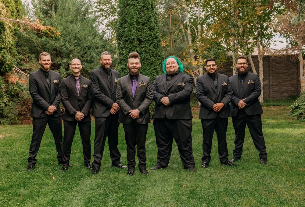 Seven men in black suits posing outdoors on green grass with lush garden backdrop for a formal group photo.