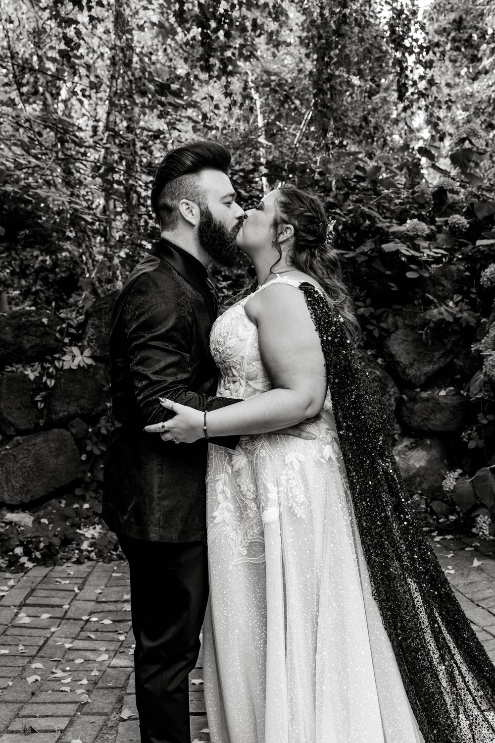 Black and white photo of a bride and groom sharing a kiss outdoors, surrounded by lush greenery and a stone wall.