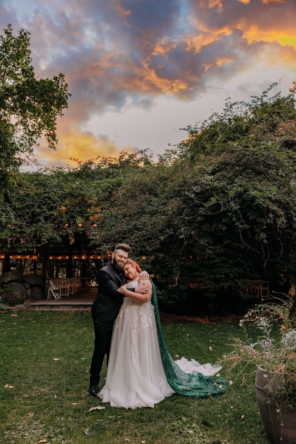 Newlyweds embrace at sunset in garden venue, bride wearing teal cape wedding gown.