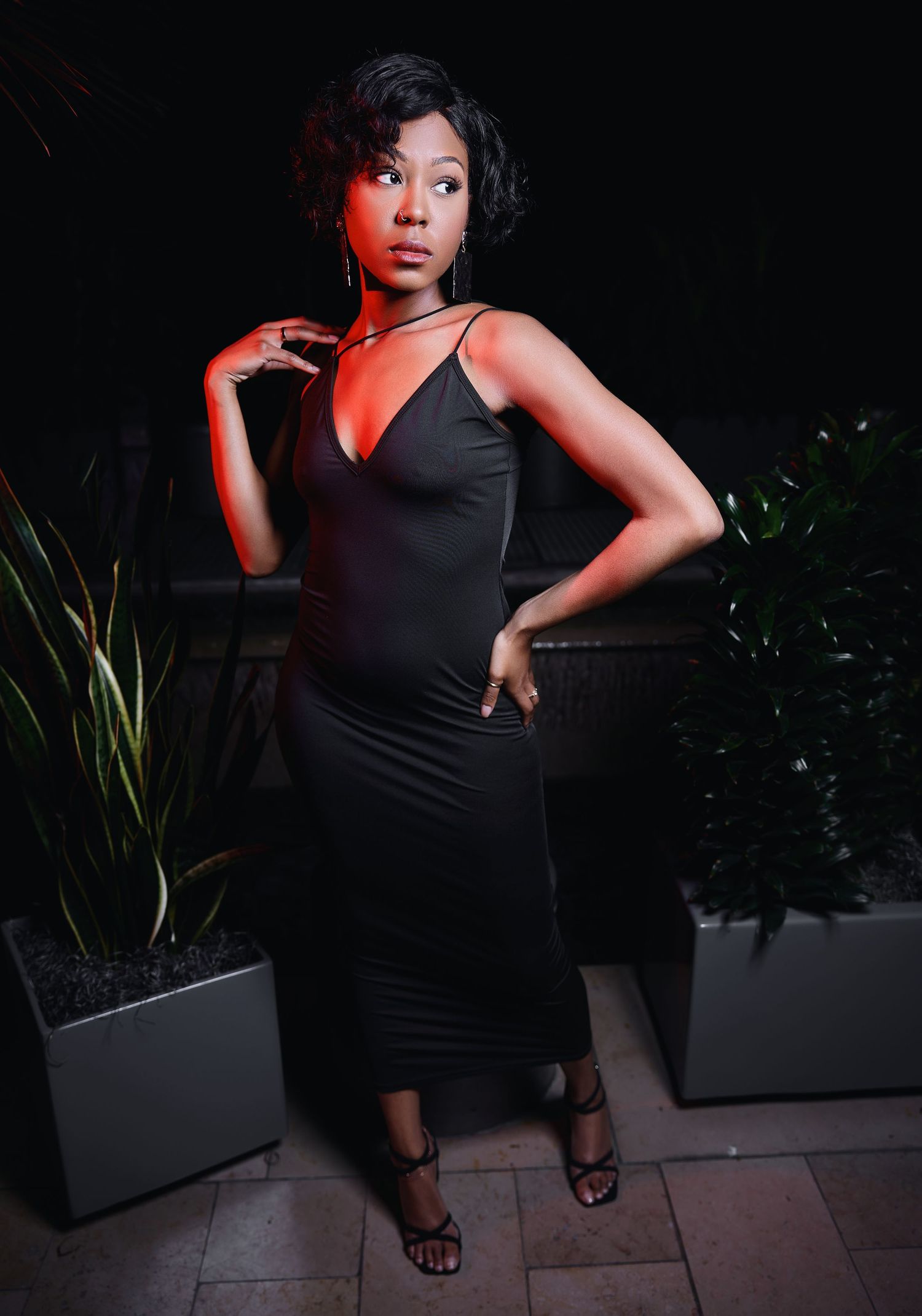 Dramatic portrait of woman in black dress posing against dark background with plants.