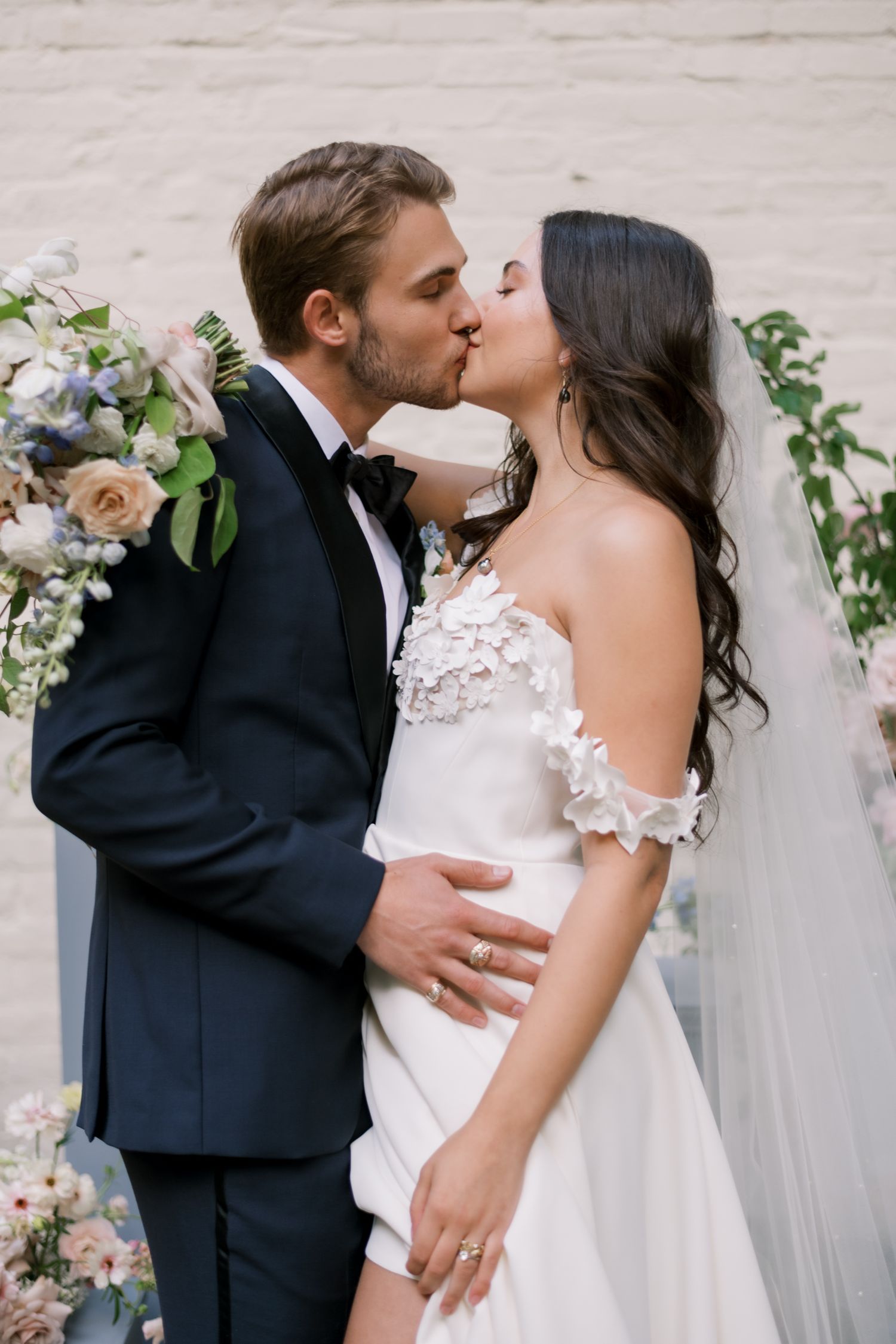 Elegant bridal portrait with flowing white veil adorned with floral appliques against a red curtain backdrop.